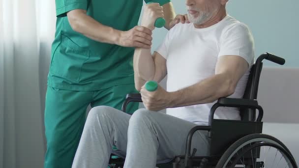 Uomo anziano in sedia a rotelle braccia flettenti con manubri, assistito da infermiera, riabilitazione — Video Stock
