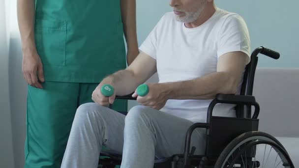Uomo anziano seduto sulla sedia a rotelle, con i manubri in mano, che parla con l'infermiera, recupero — Video Stock
