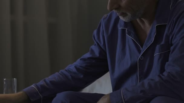Стареющий мужчина сидит на краю кровати в темноте, принимает лекарства, снотворное — стоковое видео