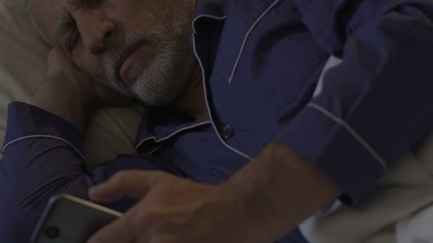 Aged man lying in bed awake at night scrolling mobile phone, problems with sleep — Stock Video