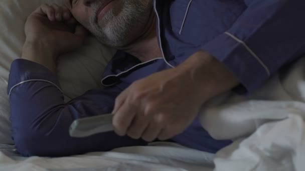 Hombre mayor acostado por la noche incapaz de dormir, cambiando canales de televisión con control remoto — Vídeos de Stock