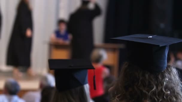 Estudiantes felices con sombreros y vestidos viendo la ceremonia de graduación, recibiendo el diploma — Vídeos de Stock