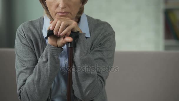 Old woman sitting on couch, holding hands on walking stick, looking sad, grief — Stock Video