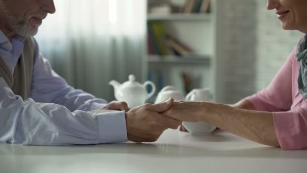 Старша пара сидить за столом з чашкою чаю, чоловік бере жінку за руку, щастя — стокове відео