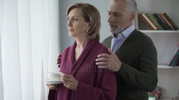 Senior kvinna som står med kopp te av fönster, man kramar henne bakifrån — Stockvideo
