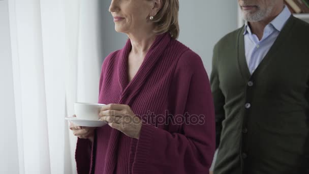 Ältere Frau mit einer Tasse Tee am Fenster, Mann umarmt sich dahinter, küsst sich auf die Wange — Stockvideo