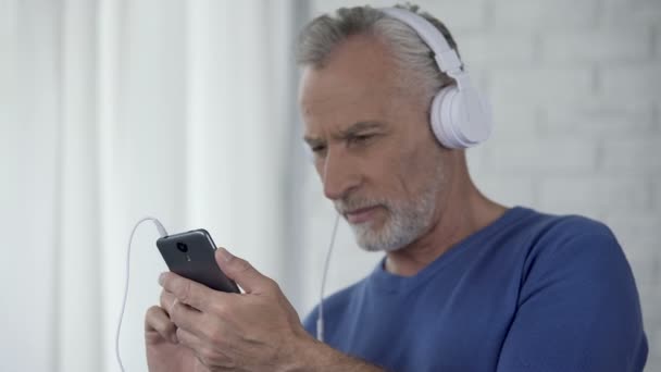 Homem sênior na tela de rolagem de fones de ouvido do smartphone, aplicativo fácil de usar — Vídeo de Stock