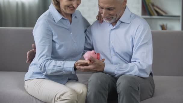 Retired couple on couch holding piggy bank, matured paid out insurance, secure — Stock Video