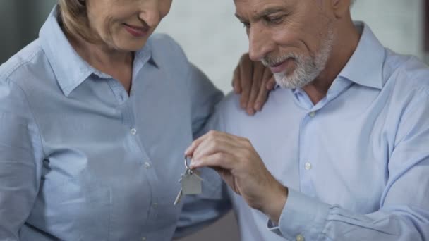 Senior paret sitter på soffan, mannen håller upp hus nycklar, fastigheter köper — Stockvideo