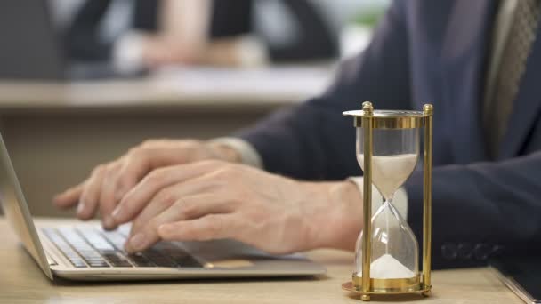 Homme en costume travaillant sur ordinateur portable au bureau, sablier debout sur la table, le temps — Video