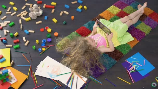 长头发的小女孩躺在五颜六色的地毯上, 在平板电脑上玩游戏 — 图库视频影像