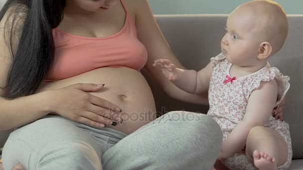 Дитяча дівчинка і вагітна жінка сидять на дивані, дитина злегка б'ється в живіт — стокове відео