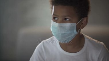 Lonely sick Afro-American boy sitting on couch in face mask, epidemic quarantine
