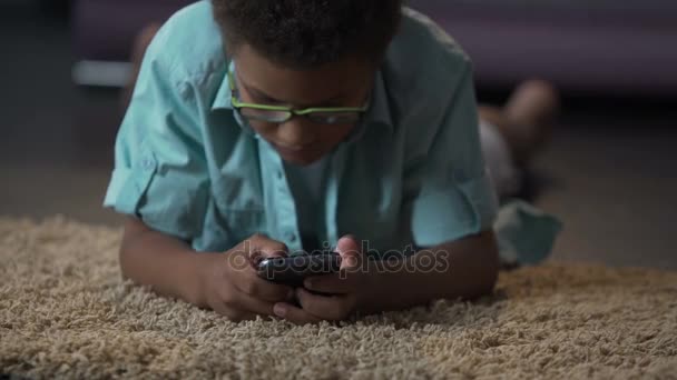 Criança afro-americana jogando em aplicação no telefone confortavelmente deitado no chão — Vídeo de Stock