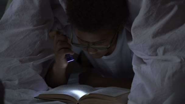 Молодий мрійник читає пригодницькі книги, що лежать під ковдрою з ліхтариком — стокове відео