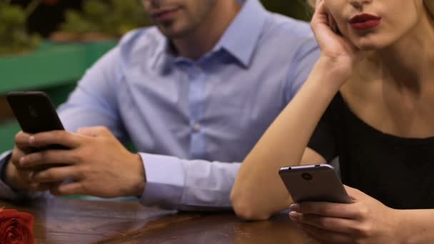 Молодая пара, сидящая за столом, поглощенная мобильными телефонами, сетевой зависимостью — стоковое видео