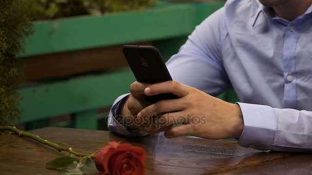 Mężczyzna siedzący przy stole za pomocą telefonów komórkowych, czerwony Róża, leżącego w przód, czekając na datę — Wideo stockowe