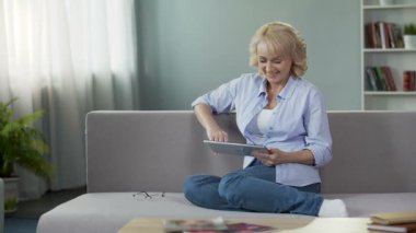 Mutlu orta yaşlı kadın evde kanepede oturan tablet, komik videoların görüntülenmesi