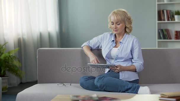 Όμορφος ξανθιά ανώτερων γυναίκα κάθεται στον καναπέ και κύλιση στο tablet, εφαρμογές — Αρχείο Βίντεο