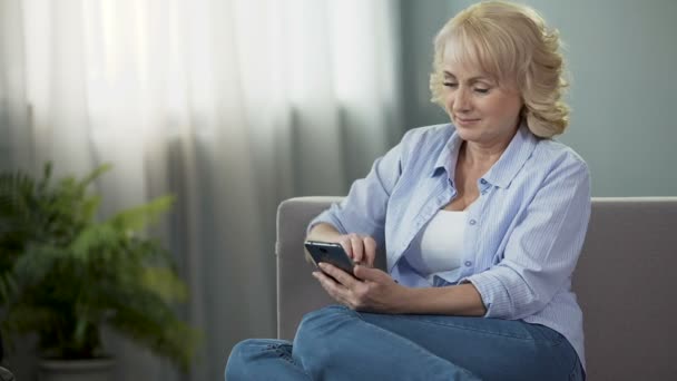 Mujer madura alegre desplazamiento noticias en las redes sociales en el teléfono inteligente en casa, aplicación — Vídeos de Stock