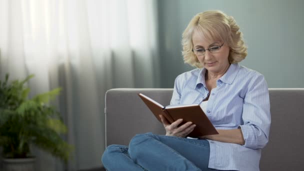Ziemlich reife Frau sitzt auf dem Sofa und liest Selbstentwicklungsbuch, Hobby — Stockvideo