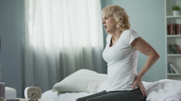 Blonde reife Dame sitzt auf dem Bett und berührt ihren Rücken, Radikulitis und Schmerzen — Stockvideo
