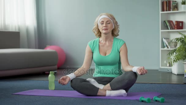 Orta yaşlı sarışın kadın pratik lotus poz evde, yoga egzersizleri yapıyor — Stok video