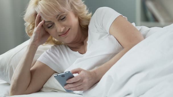 高级女性躺在床上, 手持手机和微笑, 来自心爱的信息 — 图库视频影像