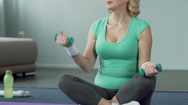 床にあぐらで座っている高齢の女性のスポーツ、ダンベルを持つ腕を屈曲 — ストック動画