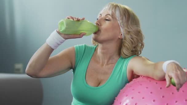 Mujer mayor apoyada en la pelota de fitness, agua potable después del entrenamiento, hidratación — Vídeos de Stock