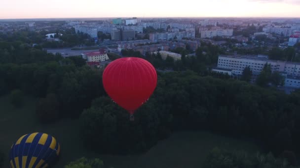 Montgolfière rouge flottant au-dessus de la zone verte de la ville à l'aube, vol précoce du festival — Video