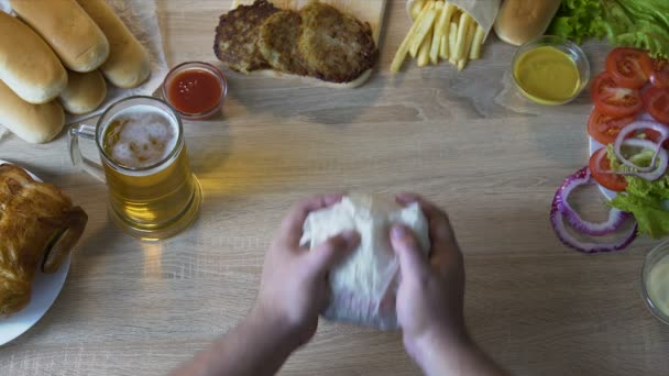 Руки толстяка распаковывают свежий бургер и несут его к его рту, жирная еда — стоковое видео