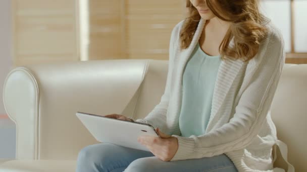 Девушка сидит на диване и выбирает новое платье на планшете, онлайн покупки — стоковое видео
