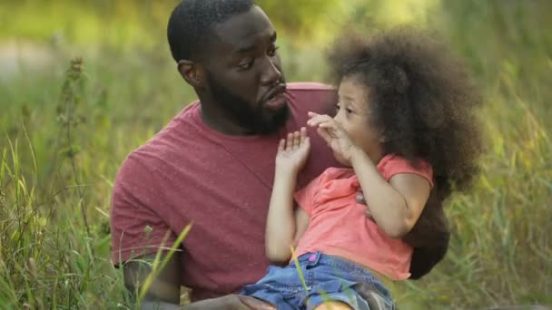 Divertido papá y su hija haciendo caras tontas juntos, sentado en medio de la hierba — Vídeos de Stock