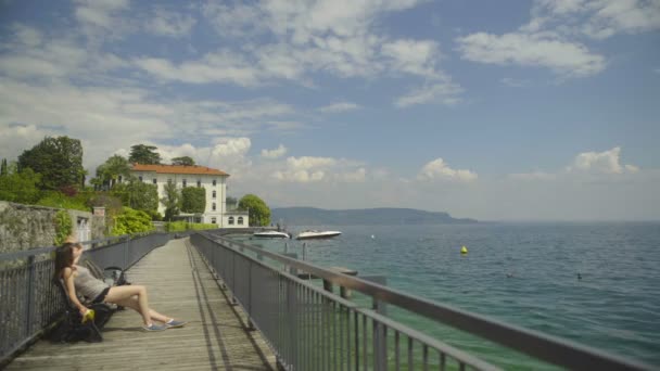 Βερόνα, Ιταλία - Circa Μαρτίου 2016: Οι τουρίστες κοντά στη λίμνη. Γυναίκα που κάθεται στον πάγκο στο pier, κάνοντας ηλιοθεραπεία και απολαμβάνοντας τη θέα στη λίμνη Γκάρντα — Αρχείο Βίντεο