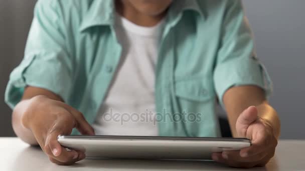 Маленький хлопчик сидить за столом і тримає планшет, граючи в комп'ютерні ігри, залежність — стокове відео