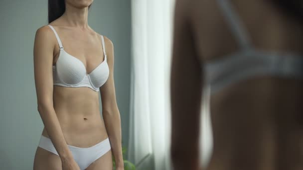 Молодая женщина смотрит в зеркало, девушка недовольна своим размером груди — стоковое видео