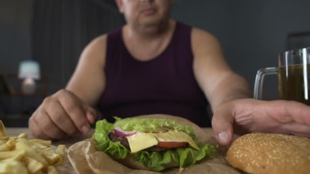 Переедающий, толстый мужчина готовит большой бургер, гурман любуется едой, крупным планом — стоковое видео