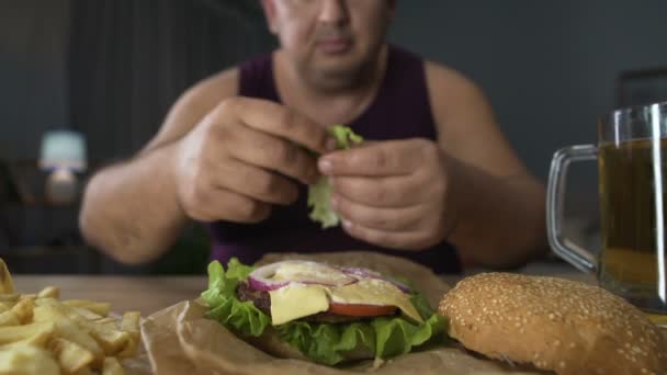 Мужчина украшает высококалорийный бургер листьями салата, готовится к еде, ожирение — стоковое видео