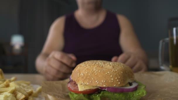 Homem com excesso de peso mordendo grande pedaço de hambúrguer, comer demais à noite, obesidade — Vídeo de Stock