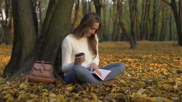 かわいい女の子公園でツリーの下に座って、コーヒーを楽しんで、彼女の日記を読む — ストック動画