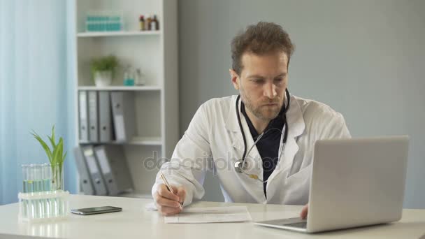 Läkaren tittar på testresultat på laptop och skriva ner patientjournaler, medicin — Stockvideo