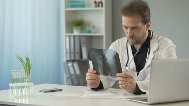 Cirurgião atento olhando para a radiografia óssea e enviando e-mail para o paciente no laptop — Vídeo de Stock