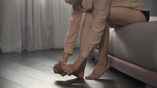 Девушка одевает колготки на тонкие ноги, сидя в спальне, собирается на работу — стоковое видео