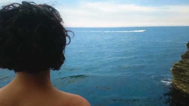 Mujer de pelo rizado de pie en el acantilado y ver yate blanco en el mar — Vídeos de Stock