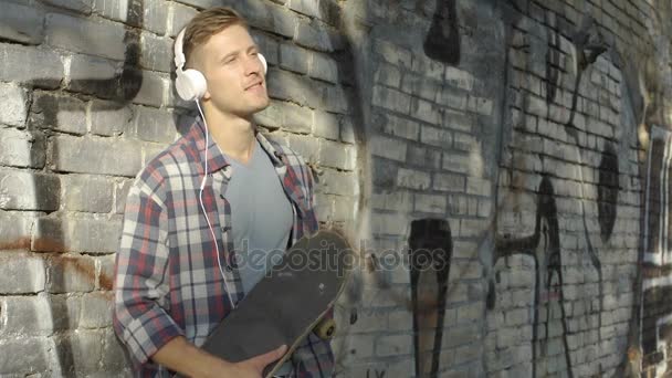 Hombre guapo con monopatín escuchando música favorita en el teléfono inteligente, hobby — Vídeos de Stock