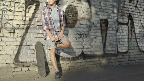 Человек с наушниками стоит у стены граффити и слушает музыку по мобильному телефону — стоковое видео