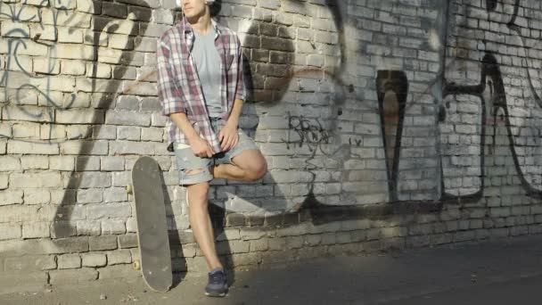 Hombre con auriculares de pie cerca de la pared de graffiti y escuchando música en el teléfono celular — Vídeos de Stock