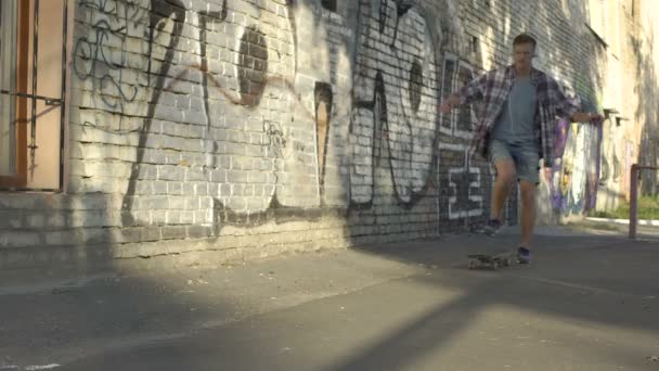 Joven vigoroso montando monopatín cerca de la pared de graffiti en la calle, hobby — Vídeos de Stock