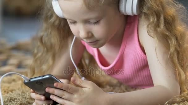 Pequeña dama acostada en la alfombra y escuchando música en auriculares en el teléfono celular — Vídeos de Stock